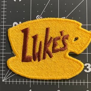 Może przedstawiać: Żółta naszywka z filcu z haftowanym brązowym tekstem "Luke's".
