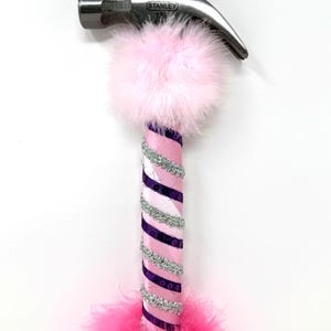 May include: A decorative hammer with a silver head and a pink and purple handle. The handle is wrapped with pink and purple ribbon, silver sequins, and pink feathers. A pink pom-pom and a gold pom-pom are attached to the hammer head.