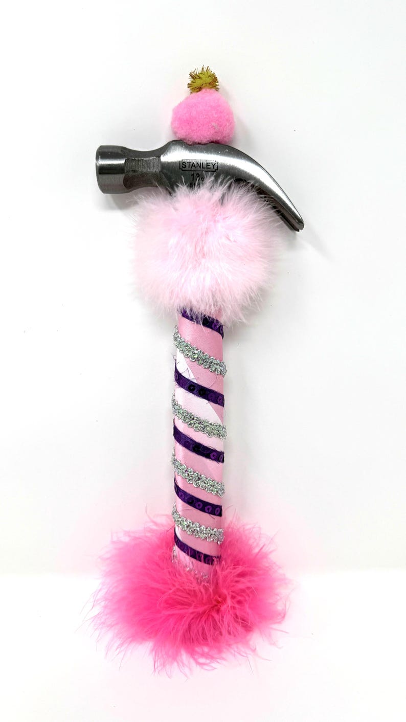 May include: A decorative hammer with a silver head and a pink and purple handle. The handle is wrapped with pink and purple ribbon, silver sequins, and pink feathers. A pink pom-pom and a gold pom-pom are attached to the hammer head.