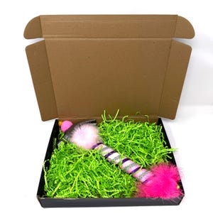 May include: A cat toy in a black box with green paper filler. The toy is pink, purple, and silver with a fluffy pink end and a pink pom-pom. The box is inside a brown cardboard box.
