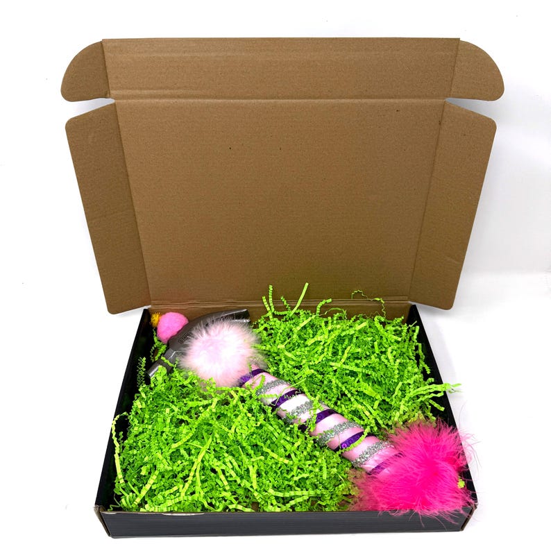May include: A cat toy in a black box with green paper filler. The toy is pink, purple, and silver with a fluffy pink end and a pink pom-pom. The box is inside a brown cardboard box.