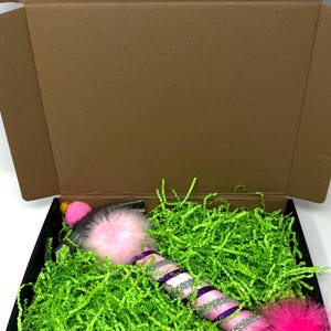 May include: A decorative wand with pink, purple, and silver accents, and fluffy pink feather ends, rests in a black box filled with green paper shreds. The wand has a pink pom-pom on one end and is packaged in a brown cardboard box.