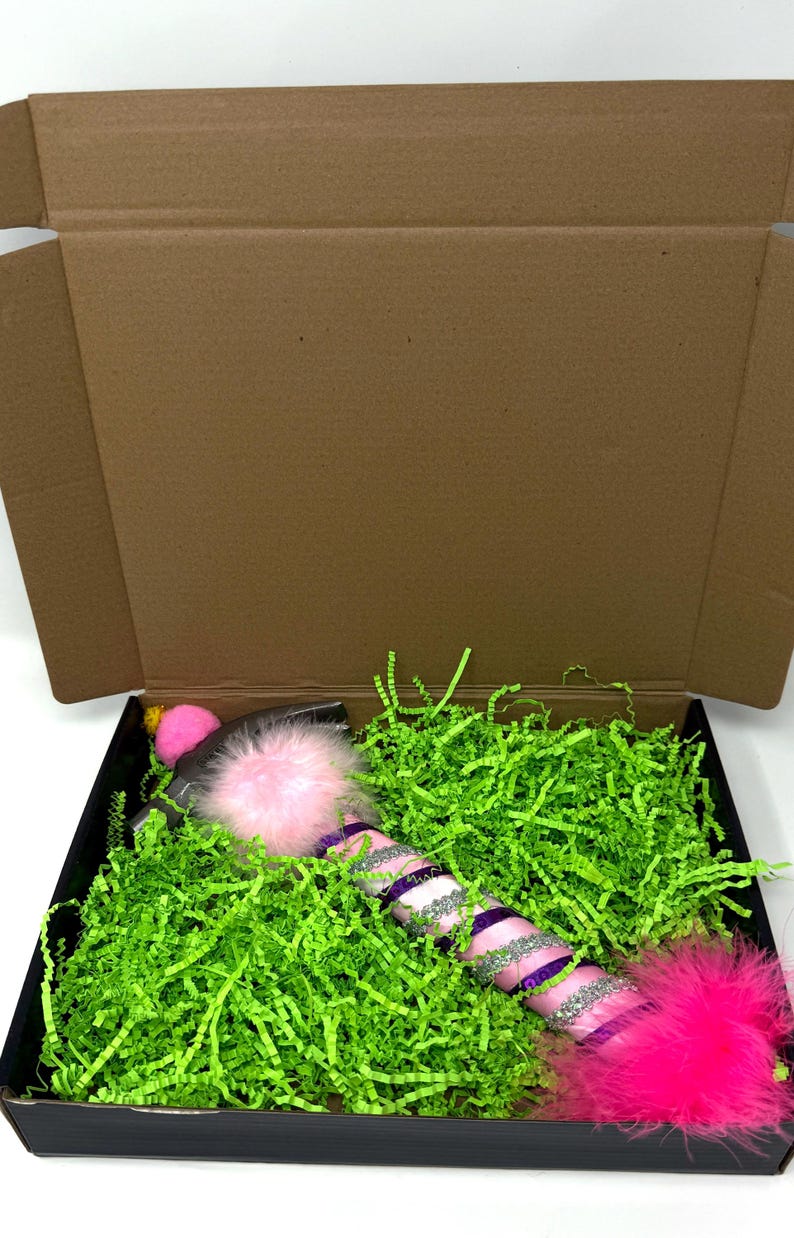 May include: A decorative wand with pink, purple, and silver accents, and fluffy pink feather ends, rests in a black box filled with green paper shreds. The wand has a pink pom-pom on one end and is packaged in a brown cardboard box.