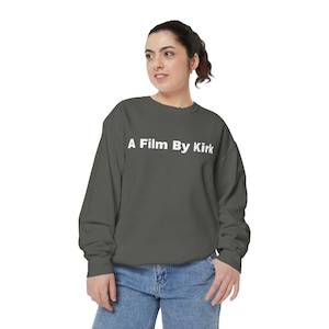 May include: A gray sweatshirt with the text "A Film By Kirk" in white. The long-sleeved crew neck sweatshirt is worn by a person wearing blue jeans. The sweatshirt is a solid color.