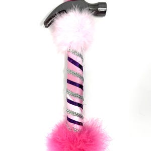 May include: A decorative hammer with a silver head and a handle wrapped in pink and purple ribbon, silver trim, and pink feathers. A small pink hat with a gold pom-pom sits on top of the hammer head. The hammer is a unique, whimsical tool.