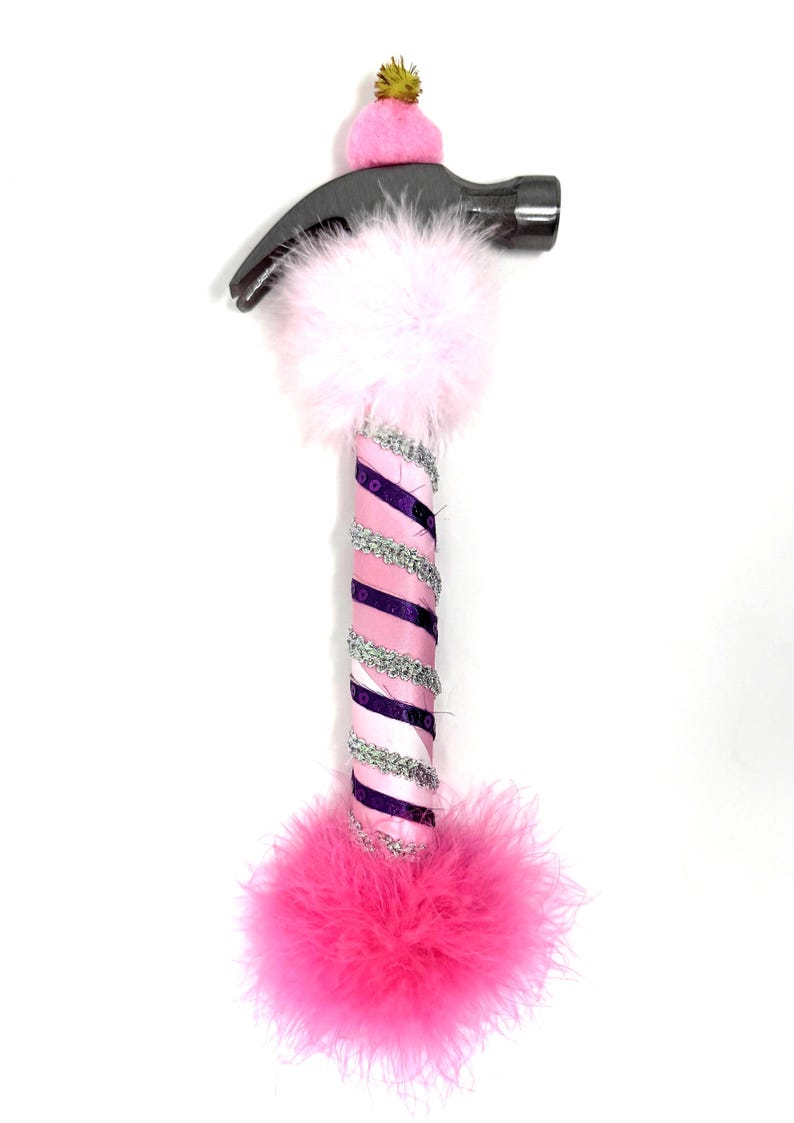 May include: A decorative hammer with a silver head and a handle wrapped in pink and purple ribbon, silver trim, and pink feathers. A small pink hat with a gold pom-pom sits on top of the hammer head. The hammer is a unique, whimsical tool.