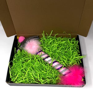 May include: A decorative hammer with a pink and purple striped handle, adorned with pink feather accents and a pink pom-pom. The hammer is presented in a black box filled with green shredded paper.