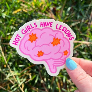 May include: A pink sticker with the text "HOT GIRLS HAVE LESIONS" in a bubble letter font. The sticker features a brain graphic with orange star-shaped accents. The sticker is held against a background of green grass.