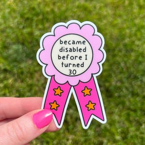 Könnte beinhalten: Ein rosa-weißer Aufkleber in Form einer Rosette mit dem Text "became disabled before I turned 30". Der Aufkleber hat zwei rosa Bänder mit orangefarbenen Sternen.