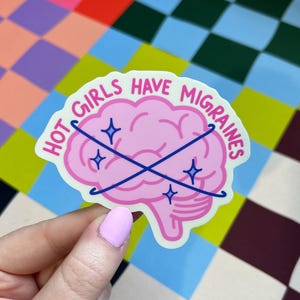 May include: A die-cut sticker featuring a pink brain illustration with two crossed dark blue lines and four small blue star icons. The text "HOT GIRLS HAVE MIGRAINES" is printed in a dark pink font along the top curve of the brain.
