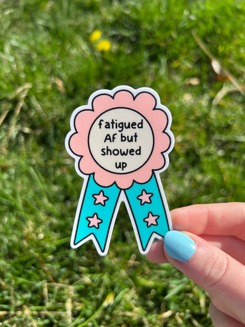 Pu&ograve; includere: Un adesivo fustellato a forma di nastro premio con un bordo rosa smerlato e un centro color crema. Il testo "fatigued AF but showed up" &egrave; scritto in nero. Due code di nastro blu brillante presentano piccole icone a forma di stella rosa.
