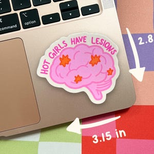 May include: A die-cut sticker featuring a pink brain illustration with orange star-shaped accents and the text "HOT GIRLS HAVE LESIONS" in pink lettering. The sticker measures 2.80 inches by 3.15 inches as indicated by the white arrows.