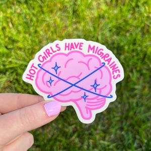 May include: A die-cut vinyl sticker featuring a pink brain illustration with the text HOT GIRLS HAVE MIGRAINES in hot pink lettering. Two crossed blue lines with small blue star accents overlay the brain, creating a graphic design on a white background.