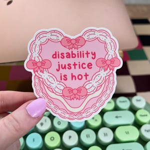 May include: A pink heart-shaped sticker with the text "disability justice is hot" in red. The sticker has a decorative border with bows. The sticker is held over a green and white keyboard.
