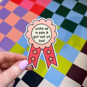 May include: A scalloped award ribbon sticker with a light pink center and red tails. The center features the black text "woke up in pain & got out of bed". The two red tails each contain two small pink star outlines, creating a decorative design.
