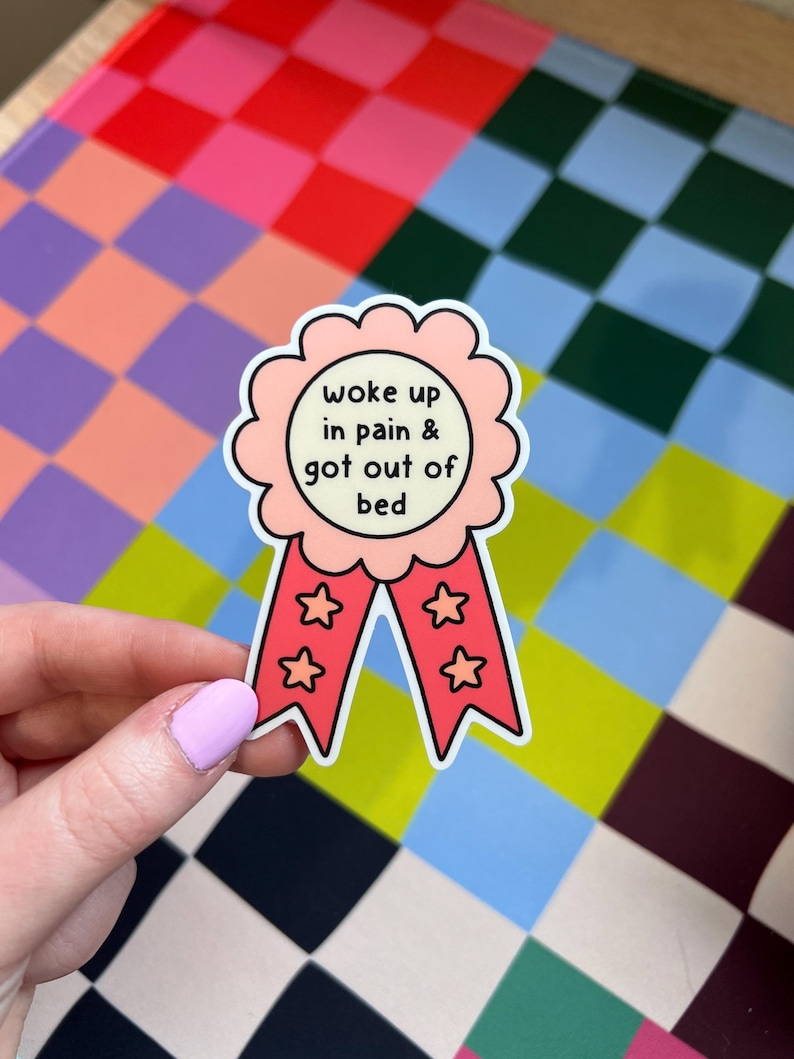May include: A scalloped award ribbon sticker with a light pink center and red tails. The center features the black text "woke up in pain & got out of bed". The two red tails each contain two small pink star outlines, creating a decorative design.