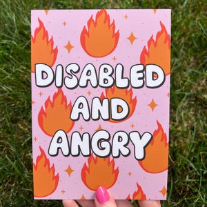 May include: A pink greeting card with the words "DISABLED AND ANGRY" in white, outlined in black. The card features orange flame illustrations and star accents. The card is held by a person with pink nail polish.