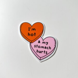 May include: Two heart-shaped stickers. The top heart is orange with the text "I'm hot" in black. The bottom heart is pink with the text "& my stomach hurts" in black. Both hearts have a white border.