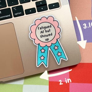 Pu&ograve; includere: Un adesivo a forma di nastro premio con un bordo smerlato rosa e code blu con stelle rosa. Il testo centrale recita "fatigued AF but showed up". L'adesivo misura 3,10 pollici di altezza e 2 pollici di larghezza, come indicato dalle frecce.