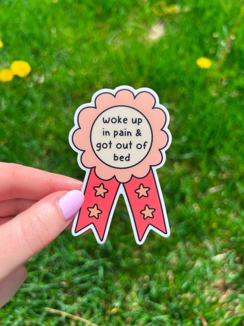 May include: A die-cut sticker shaped like a circular award ribbon with a scalloped edge. The center is cream-colored with the black text 'woke up in pain & got out of bed'. Two red ribbons hang down, each decorated with two orange star outlines.