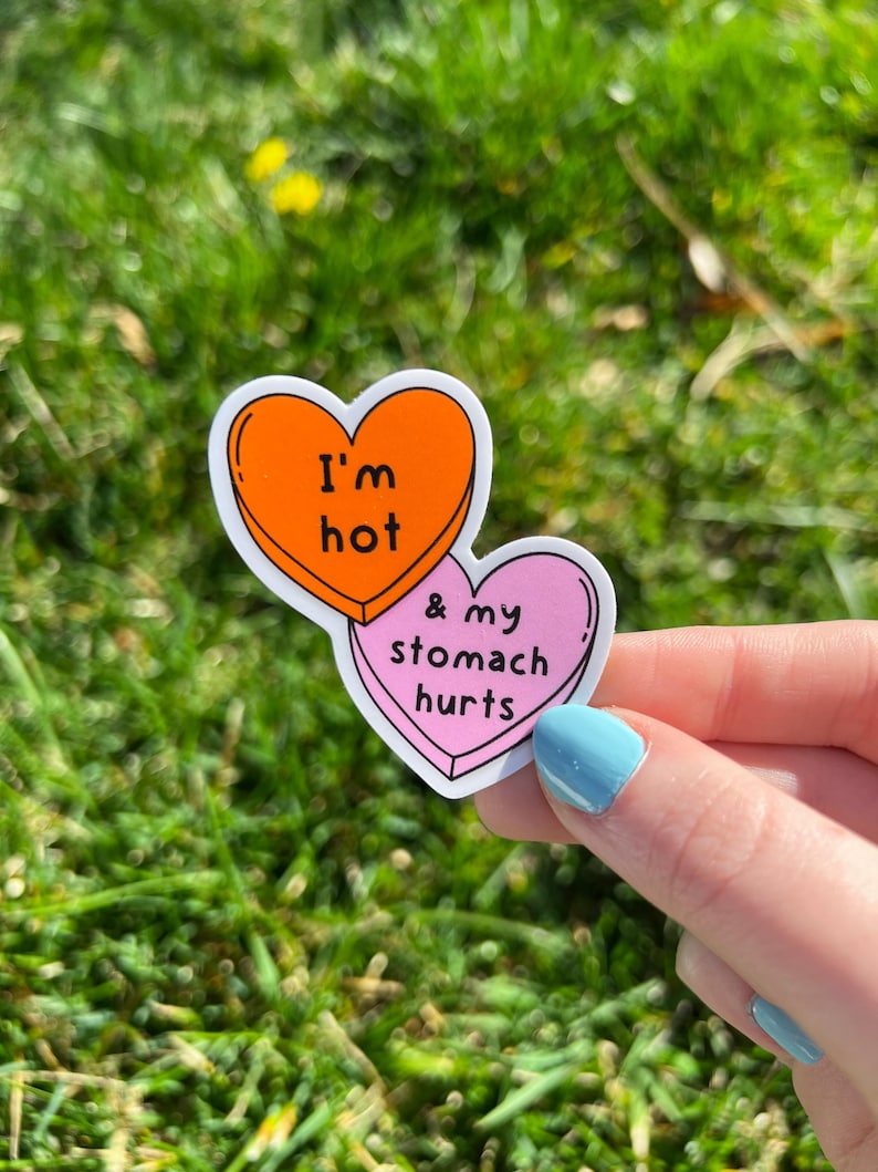 May include: Two heart-shaped stickers with text. The top sticker is orange with the words "I'm hot" in black. The bottom sticker is pink with the words "& my stomach hurts" in black. The stickers are held by a hand with blue nail polish.