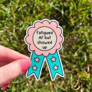 May include: A sticker with a pink and white scalloped edge, featuring the text "fatigued AF but showed up" in a black font. The sticker has teal ribbons with four white stars. The sticker is held over green grass.