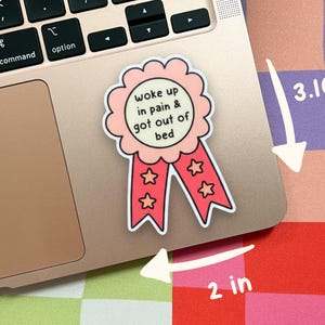 May include: A pink and red ribbon sticker with a scalloped edge and two tails featuring star icons. The center text reads 'woke up in pain & got out of bed'. The sticker measures 3.10 inches in height and 2 inches in width as indicated by arrows.