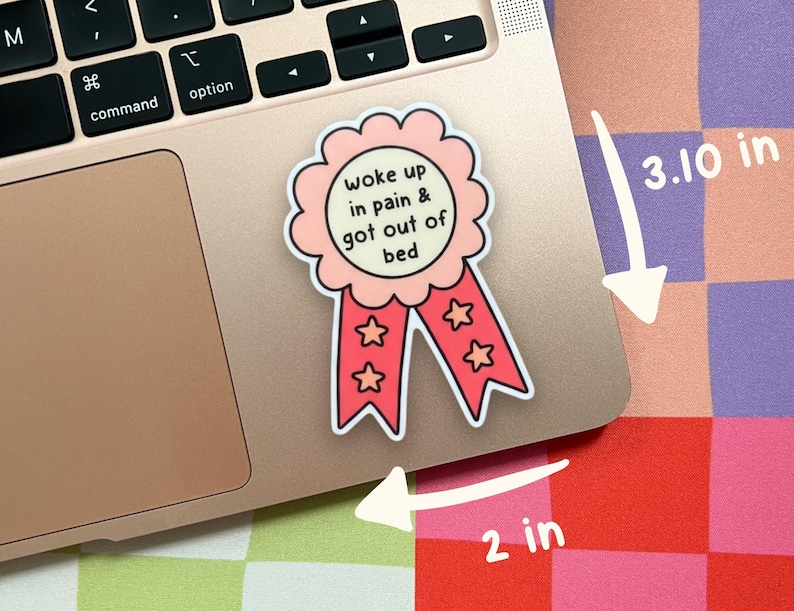 May include: A pink and red ribbon sticker with a scalloped edge and two tails featuring star icons. The center text reads 'woke up in pain & got out of bed'. The sticker measures 3.10 inches in height and 2 inches in width as indicated by arrows.