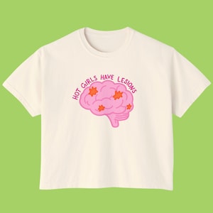 May include: A cream-colored short-sleeve t-shirt featuring a graphic of a pink brain with orange star-shaped spots. The text "HOT GIRLS HAVE LESIONS" is printed in a curved, dark pink font above the brain illustration on the front of the shirt.