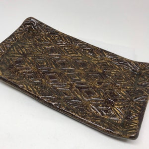 Peut inclure: Plateau rectangulaire en céramique avec un motif géométrique en losanges. Le plateau est de couleur marron foncé avec un motif marron clair. Les bords sont légèrement incurvés. Ce plateau décoratif convient au service ou à l'exposition.