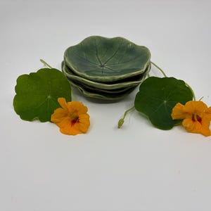 Puede incluir: Una pila de tres cuencos pequeños de cerámica en color verde salvia con acabado brillante y un patrón de nervaduras de hoja moldeado. Los cuencos están acompañados por dos flores de capuchina de color naranja brillante y dos hojas verdes.