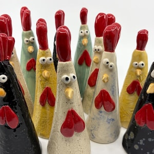May include: A group of ceramic rooster ornaments in diverse colours, including black, yellow, green, and white. Each rooster has a red comb and wattle, with white eyes and a yellow beak. The ornaments are handmade and have a whimsical design.
