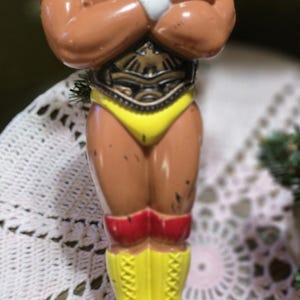 May include: A vintage Hulk Hogan action figure. The figure is brown with yellow boots, a yellow speedo, and a red tank top with "HULKAMANIA" printed on it. The figure has its arms crossed.