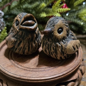 May include: Two small, brown ceramic bird figurines with detailed feather textures. The birds have open beaks and round eyes, sitting on a wooden, tiered stand. The background includes green foliage, suggesting a nature-inspired theme.