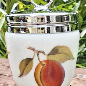 May include: A white ceramic jar with a polished silver-tone metal lid featuring a circular handle. The body of the jar is decorated with a botanical illustration of a single peach with green leaves on a branch, rendered in warm orange and red tones.