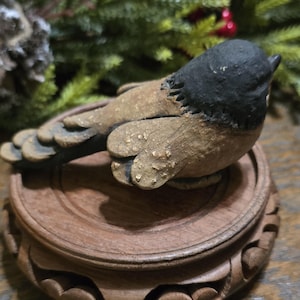 May include: A handcrafted bird figurine with a black head and brown body, perched on a decorative wooden stand. The bird has textured details and is set against a blurred background of greenery.