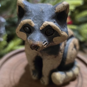 May include: A ceramic raccoon figurine with a black and tan color scheme. The raccoon is sitting on a wooden base. The figurine has detailed features, including a black mask around its eyes and striped tail.