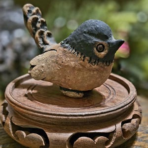 May include: A handcrafted ceramic bird figurine with a black head and a brown body, perched on a decorative wooden stand. The bird has detailed features, including a small beak and eye. The stand has carved accents.