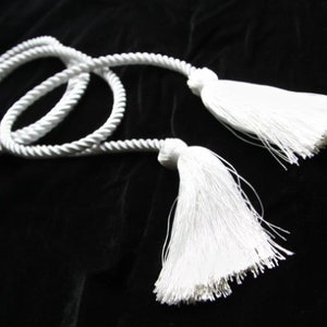 May include: White rope with two white tassels at the ends. The rope is coiled and the tassels are hanging down.