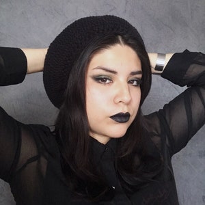 May include: A woman with dark hair and dark eye makeup is wearing a black beret and a black sheer blouse. She is looking at the camera.