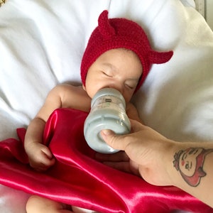 May include: A baby wearing a red devil horn hat, being fed from a baby bottle. The baby is wrapped in a red satin blanket, against a white sheet background.