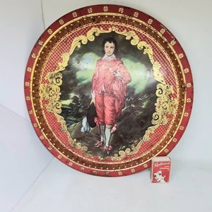 May include: A round, red metal tray with a gold rim and a gold and white design. The centre of the tray features a painting of a man in a red and white outfit standing in a landscape.