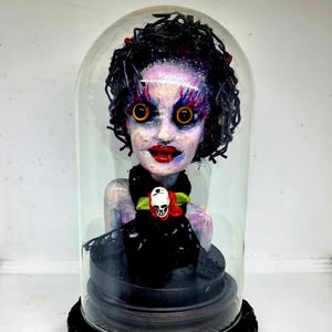 May include: A gothic doll sculpture displayed under a glass dome. The doll features a pale complexion, red lips, large yellow eyes, and dark curly hair. It is dressed in black and wears a skull pendant. The dome rests on a decorative black base.