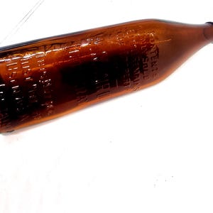 May include: A brown glass bottle with embossed text. The bottle has a long neck and a rounded body. The text appears to be a brand name and patent information. The bottle is lying on a white surface.