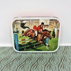 May include: Rectangular tin featuring a painted equestrian scene. A rider in a red jacket jumps a fence on a brown horse. The background shows a crowd and a green field. The tin has a white border.