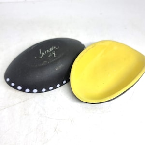 May include: Two ceramic dishes. One is black with white dots along the edge and the handwritten text "Junda - 18". The other dish is yellow. Both dishes are oval-shaped.