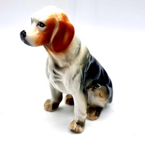 May include: A ceramic figurine of a sitting hound dog with a white chest and legs, a black back, and tan markings on the head and ears. The dog has a black nose and dark eyes, with a glossy finish and a smooth, sculpted posture facing left.