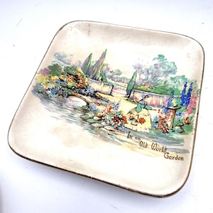 May include: A square ceramic trinket dish with a cream-coloured base and a crackled glaze finish. The centre features a colourful illustration of a garden scene with a person in a yellow skirt, a stone bridge, and the text In an Old World Garden.