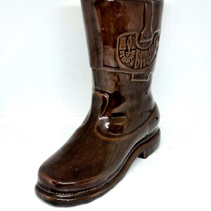 May include: A brown ceramic boot-shaped vase with a glossy finish. The vase features embossed details, including the word "Budweiser". The boot design includes a heel, sole, and a tall shaft. A unique decorative item.