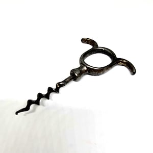 May include: Vintage metal corkscrew with a dark, aged finish. The corkscrew has a spiral worm and a handle with a circular opening and curved arms. A classic wine accessory.